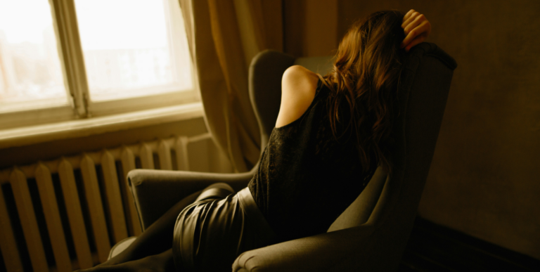 A woman sitting alone in an armchair by a window, her head resting in her hand
