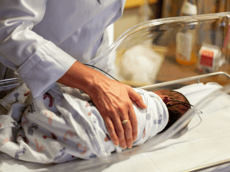 A newborn baby swaddled in a hospital cot, with a nurse's hand resting gently on them